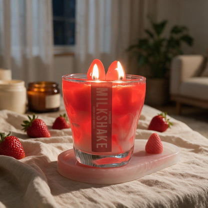 A red wax candle in a clear glass holder with two lit wicks, placed on a pink tray on a bed in a cozy bedroom setting. The candle has a vertical label reading 'MILKSHAKE'.
